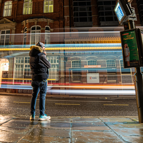 Man standing at bus stop at night. Long exposure with bus lighttrail behind him.